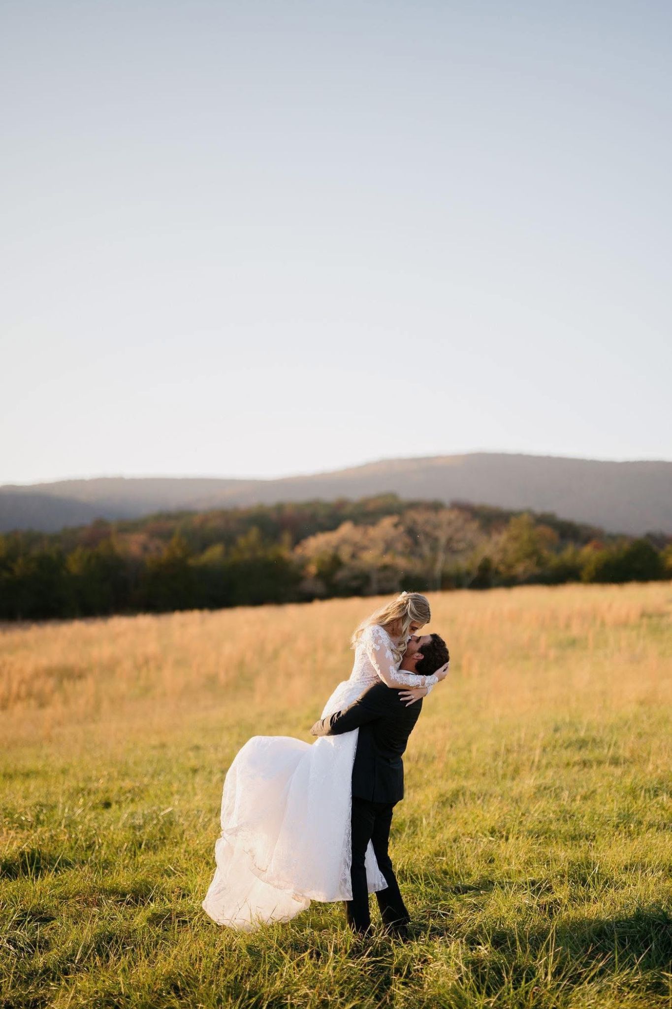 Groom holding up bride in glowing orange fall fields with long grass and Blue Ridge Mountains in the background at Canaan Springs, a rustic farm wedding venue in Northern Virginia