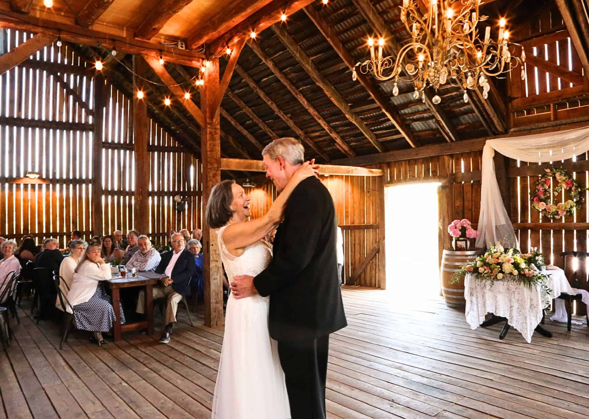 Older couple sharing their just married first dance in the barn at Canaan Springs with warm string lighting, floral arrangements, lacey tablecloth, and chandelier, a Northern VA area barn wedding venue near Winchester, VA and the Winchester area