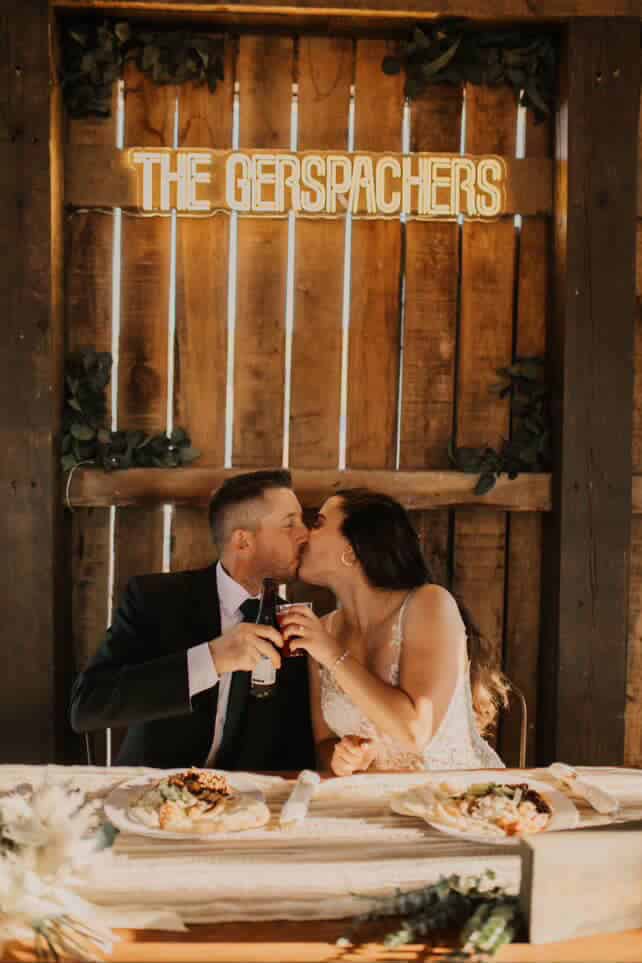 Newlywed couple kissing at their sweetheart table in the Canaan Springs barn after toasts, with a glowing sign and rustic wooden backdrop at this Northern Virginia farm wedding venue