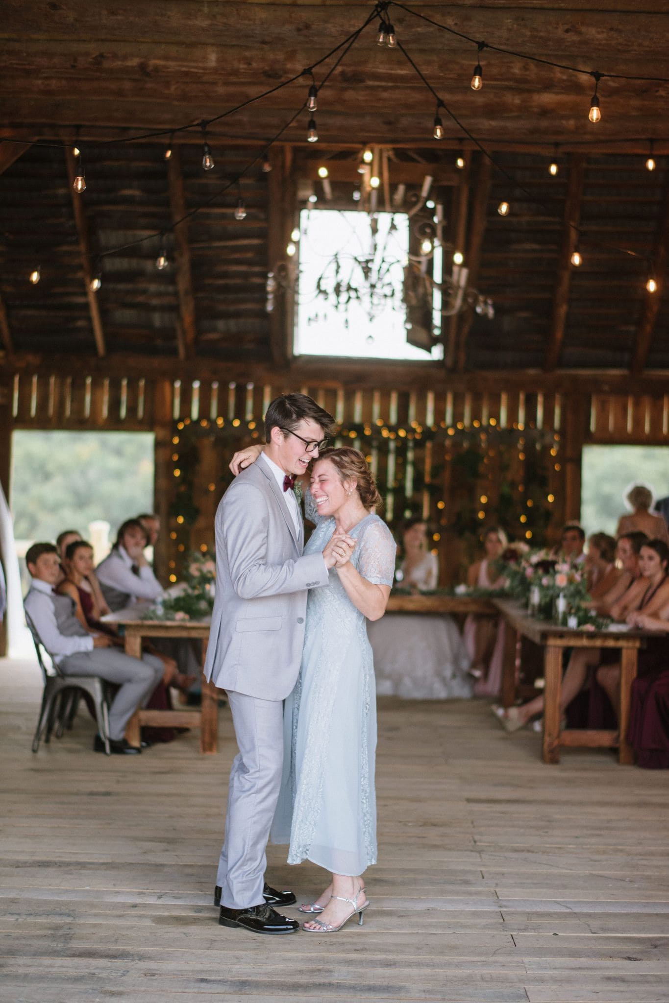 Mother and son sharing a dance in the brightly lit barn at Canaan Springs, with the wedding party seated at a large table enjoying the scenic view at this Northern Virginia farm wedding venue